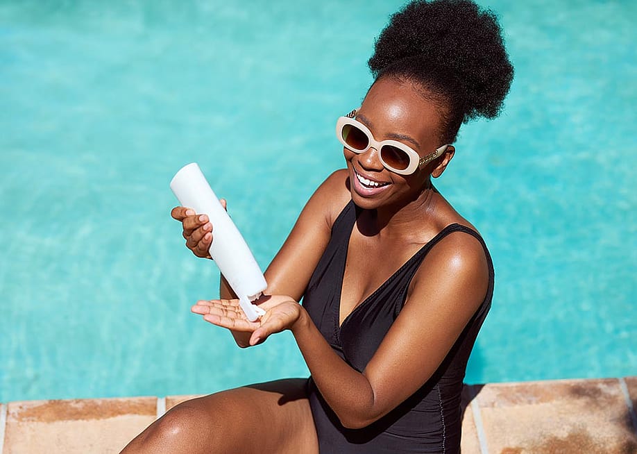 Black woman wearing black suit and white-framed sunglasses applies sunscreen sitting by pool in summer sun — Meeko Media // Shutterstock