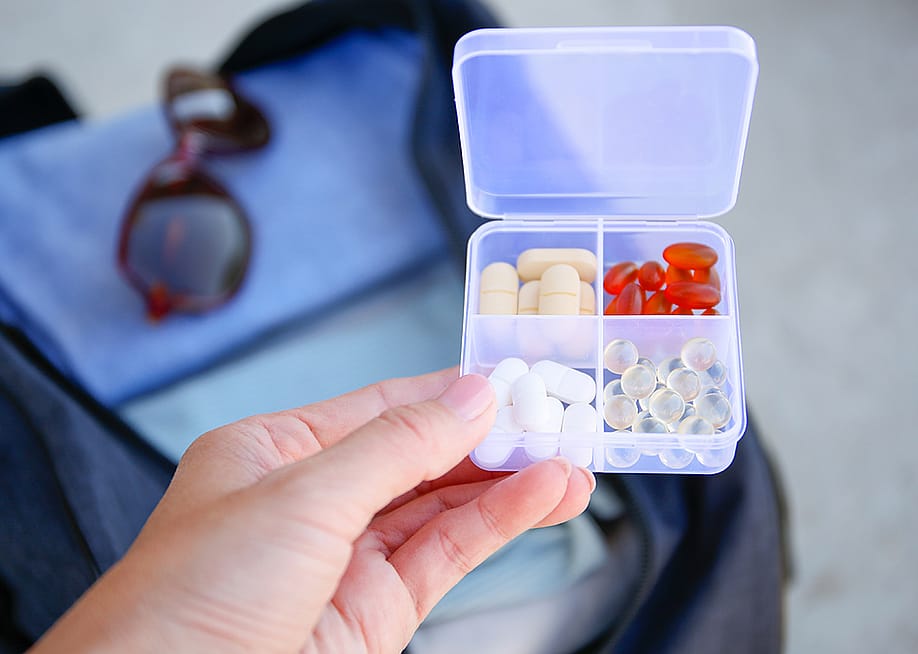 A box of pills with a travel bag in the background. — Victoria1 // Shutterstock
