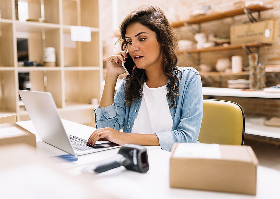 A businesswoman speaking on the phone while using a laptop in her small business production area. — JLco Julia Amaral // Shutterstock