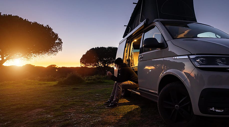 Silhouetted digital nomad sitting in his white transporter camping van, backlit by the setting sun near the California coast. — Audio und werbung // Shutterstock