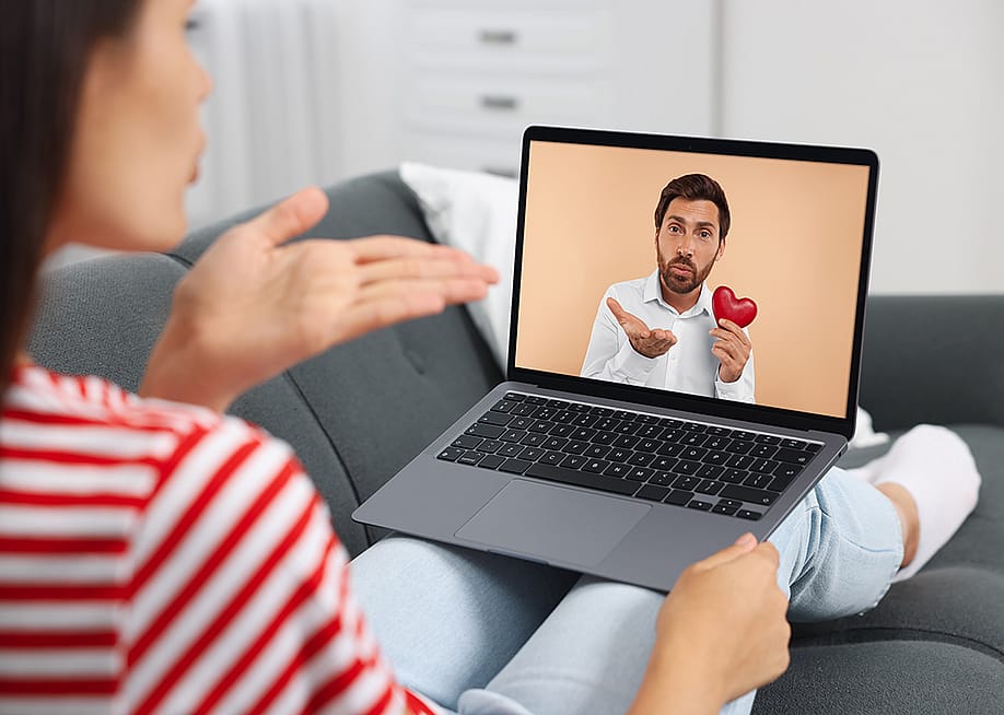 Concept of romance scam illustrated by a couple communicating long-distance via video chat, with man holding a heart figure and blowing a kiss. — New Africa // Shutterstock