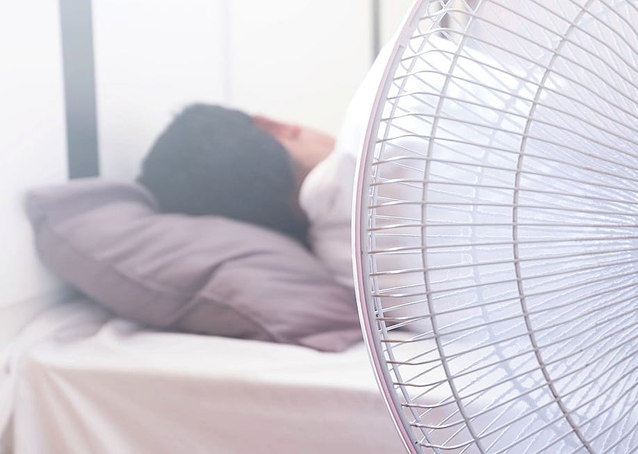 A white electric fan in the foreground in front of a person sleeping in bed, facing away. — phungatanee // Shutterstock