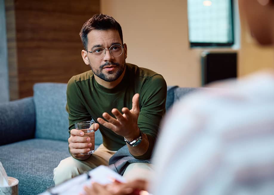 Person wearing long sleeve forest green top and glasses sits on a couch holding a glass of water and gesturing to a person with their back to their camera. — Drazen Zigic // Shutterstock.