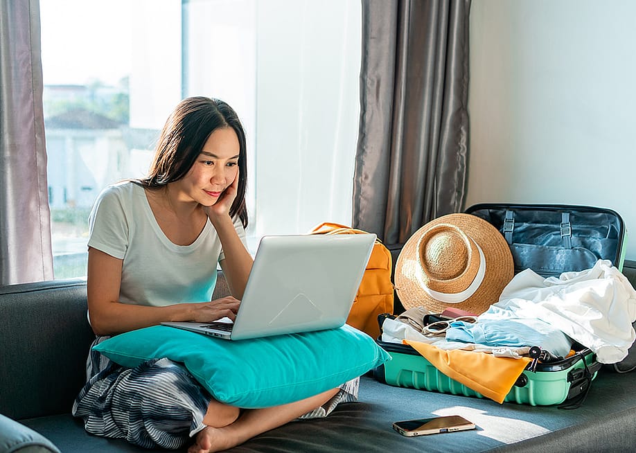 Young woman using a laptop beside an unpacked suitcase preparing for a trip. — Opat Suvi // Shutterstock