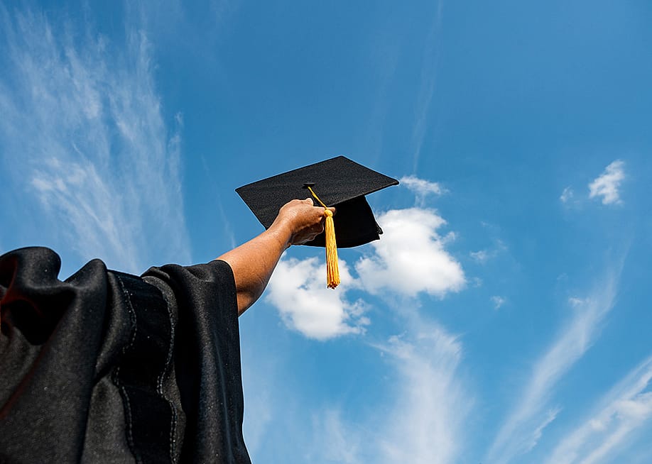 A graduate student holding up a graduation cap in the sky. — Nirat.pix // Shutterstock
