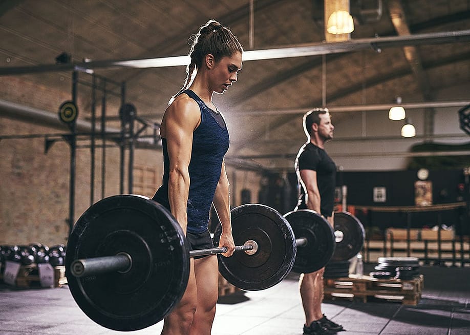 Strong woman and in foreground lifting bar bell with weights with man in background gym. — Ground Picture // Shutterstock