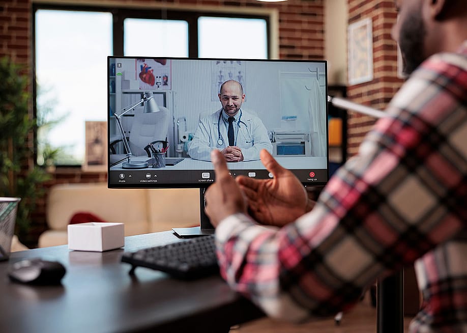Computer screen showing telehealth call with male balding doctor sitting in office with stethoscope around his neck, while person dressed in plaid long sleeve shirt in foreground converses on video call with his back to the camera. — DC Studio // Shutterstock