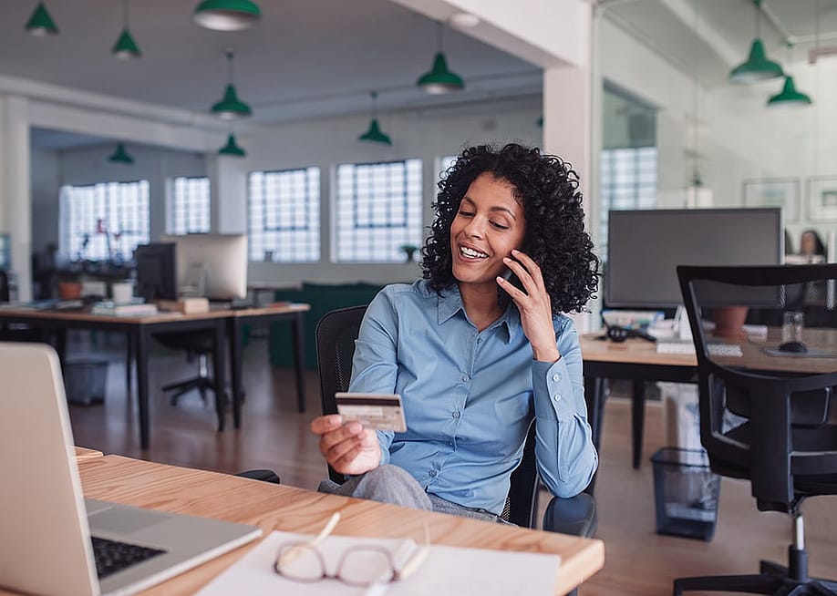 Businesswoman in her workplace looking at her credit card while on the phone. — mavo // Shutterstock.