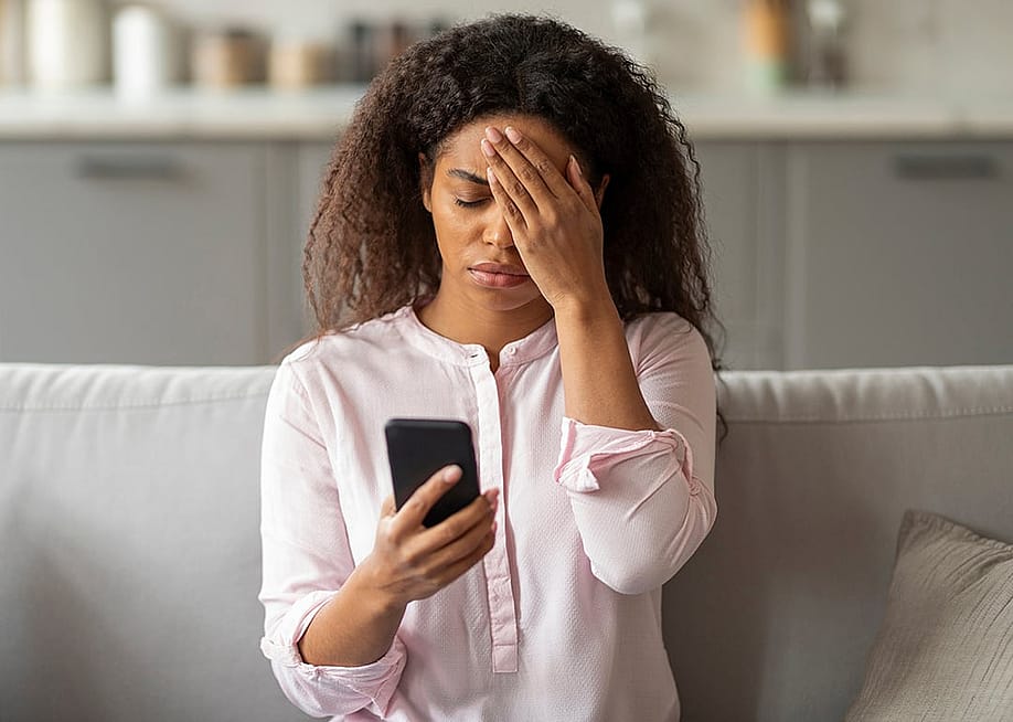A photo capturing the tension of a young African American woman sitting on her couch, visibly overwhelmed as she looks at her smartphone in a neatly arranged living room. — Prostock-studio // Shutterstock
