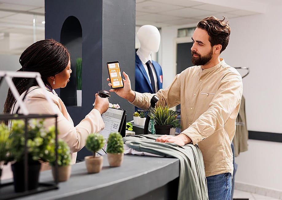 Male customer holding up smartphone with rewards coupon to be scanned by business worker at a retail shop. — DC Studio // Shutterstock