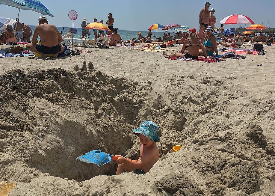 Child wearing a blue hat digging a giant hole in sand at the beach that is higher than him on both sides. — OMfotovideocontent // Shutterstock