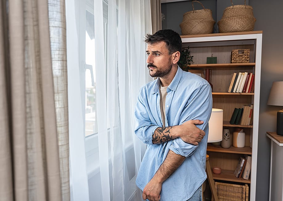 Young sad and depressed anxious looking man wearing a blue button up shirt looking through white curtains out the window. — Srdjan Randjelovic // Shutterstock