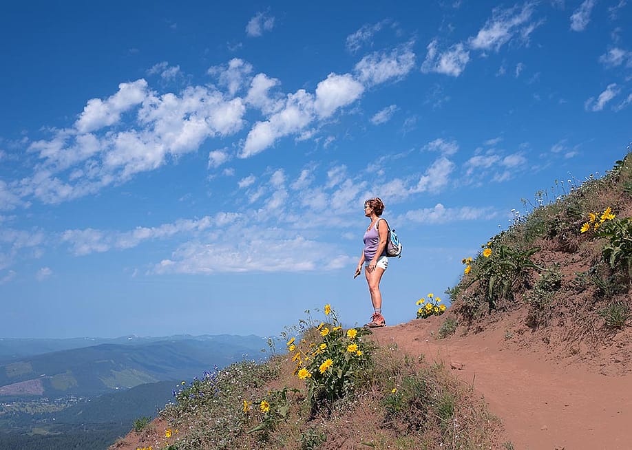 Woman looks out toward view on dirt mountain trail with yellow flowers, hiking in summer with sky and clouds in background near Portland, Oregon. — Marina Poushkina // Shutterstock