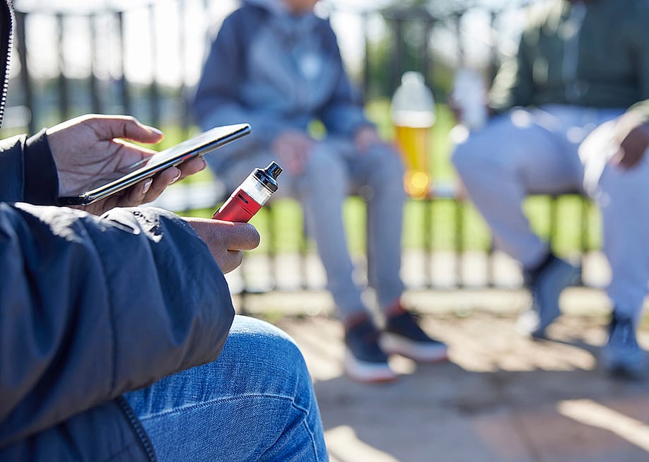 Teenager holding a vape while scrolling on the phone at the park. — Daisy Daisy // Shutterstock