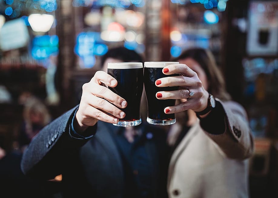 Two people holding up a pint each of stout beers to cheers, which obscures their faces from the camera in a bar. — jjmtphotography // Shutterstock