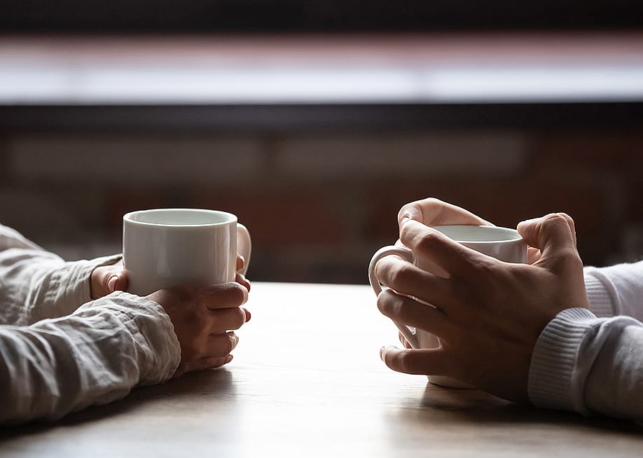 A Gen Z and Millenial sitting across each other at a table drinking coffee. — fizkes // Shutterstock