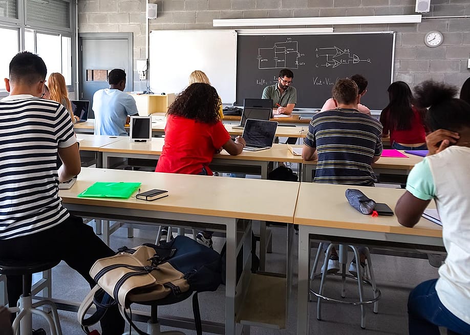 View from back of classroom of multiracial high school students in class using laptops while teacher marks exams. — Daniel Hoz // Shutterstock
