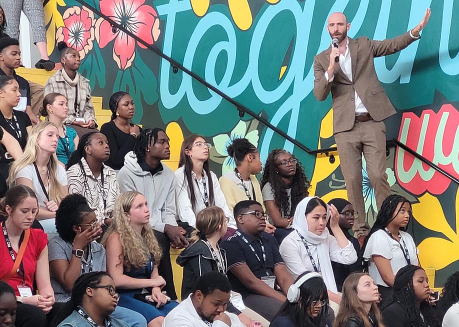 Side view of rows of high school students sitting on bleachers listening to a speaker wearing a tan suit talking into a mic. — Patrick O’Donnell for The 74