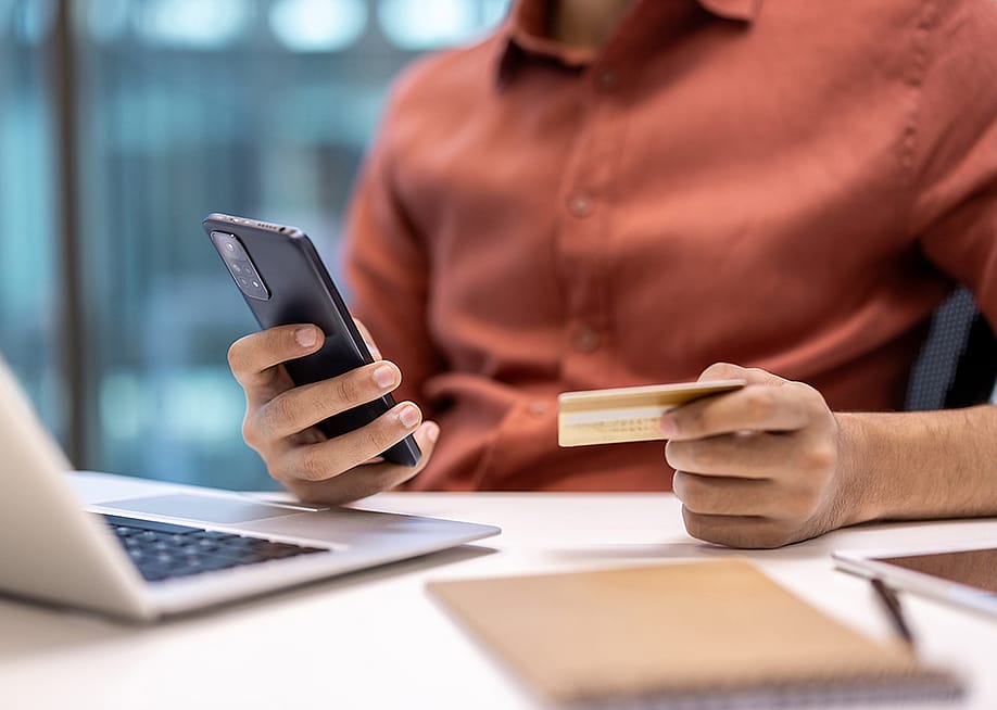 Businessperson holding a credit card while using a phone and laptop at an office desk. — voronaman // Shutterstock