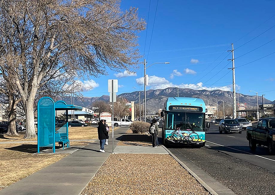 Bus riders board a blue bus in Albuquerque, next to a turquoise metal bus stop and with mountains and cloud in distance. — Erin Rode for Next City