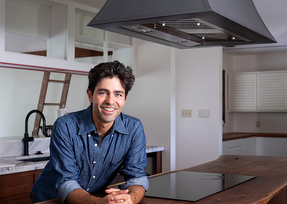 Adrian Grenier stands beneath his island-mounted CopperSmith® range hood, featuring a smooth bronze finish, paired with a handcrafted wood countertop for a warm, natural aesthetic. — World CopperSmith