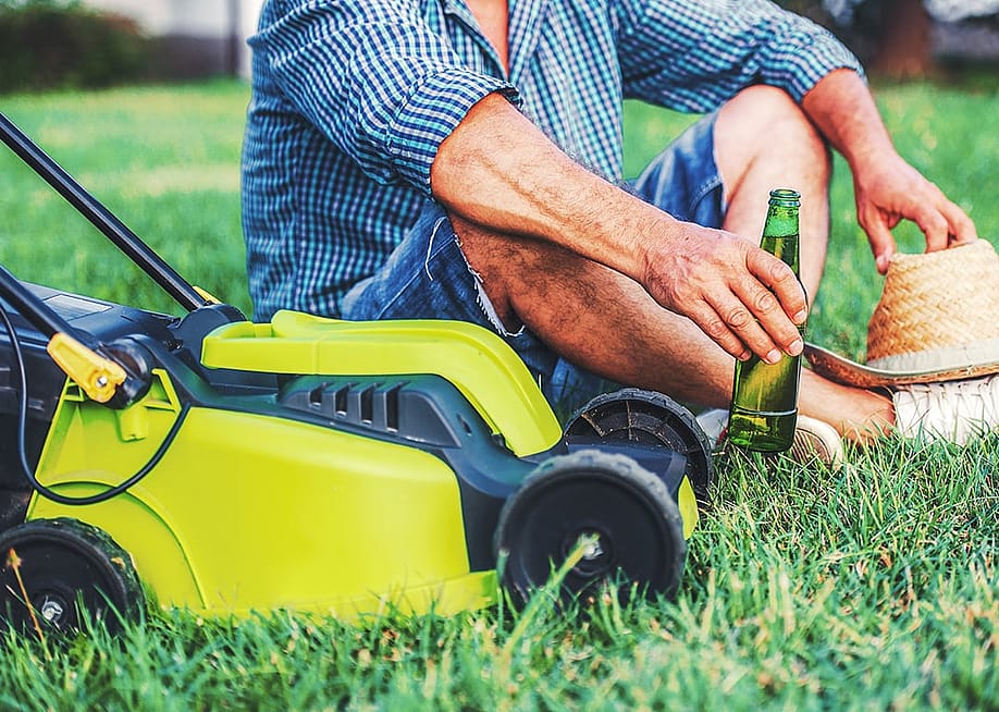 Senior man enjoying a bottle of beer beside a mower in his lawn. — Bobex-73 // Shutterstock