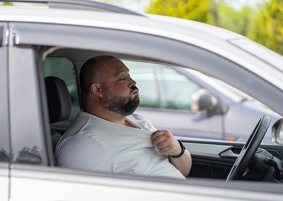 A bald driver with closed eyes seen through the passenger side window pulls at the collar of a white t-shirt and exhales. — DimaBerlin // Shutterstock