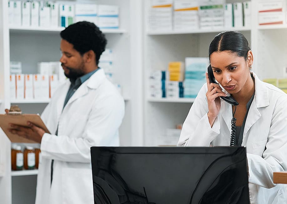 Two pharmacy employees work behind the counter in a pharmacy, one on the phone and looking at a computer and the other writes on a clipboard. — Yuri A // Shutterstock