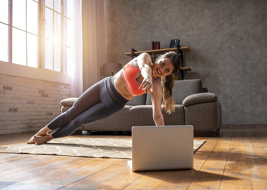 Woman doing a side plank pose on a mat on a wooden floor, following a video on a laptop, with sunlight coming in through large window in the background. — alphaspirit.it // Shutterstock