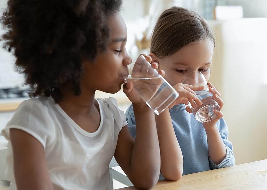 Two children, one Black with an afro and one light skinned child with long hair tied back sit at a table drinking glasses of water. — fizkes // Shutterstock