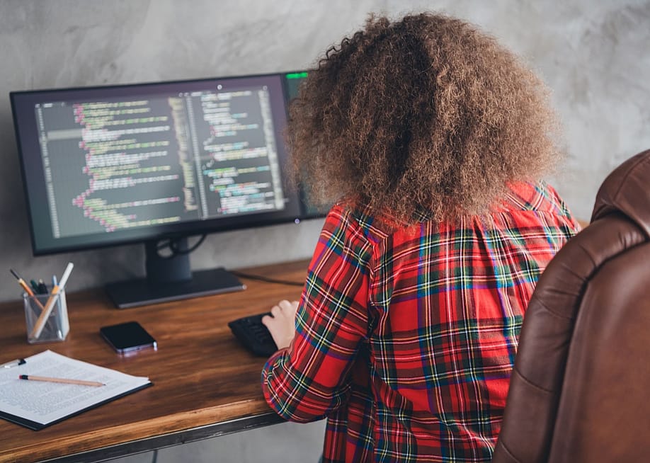 Rear view of a woman with curly hair working at a computer desk displaying code in a casual office setup wearing a checked shirt. — Roman Samborskyi // Shutterstock