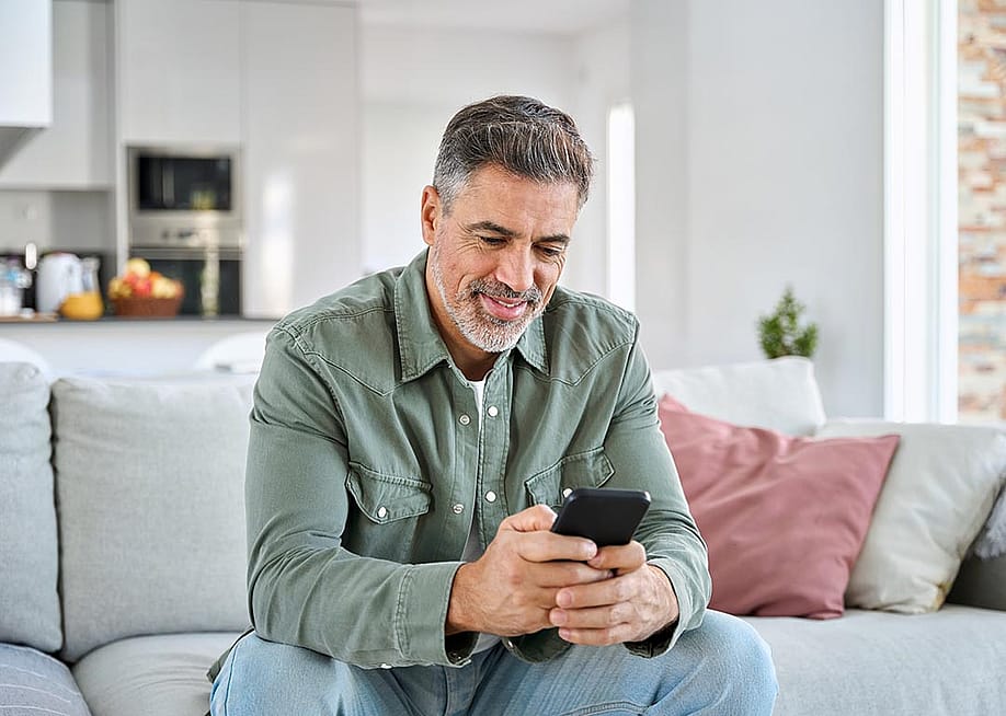 Middle aged old man using smartphone sitting on a couch with kitchen in background. — insta_photos // Shutterstock
