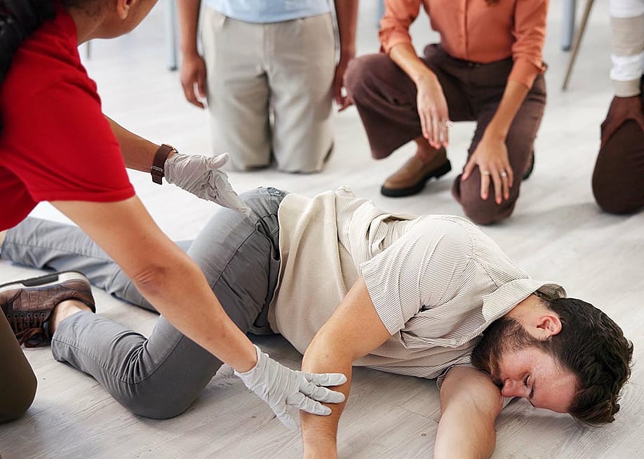 Demonstration by person teaching class for first aid, with someone as victim lying on their side and three people kneeling and watching. — Yuri A // Shutterstock