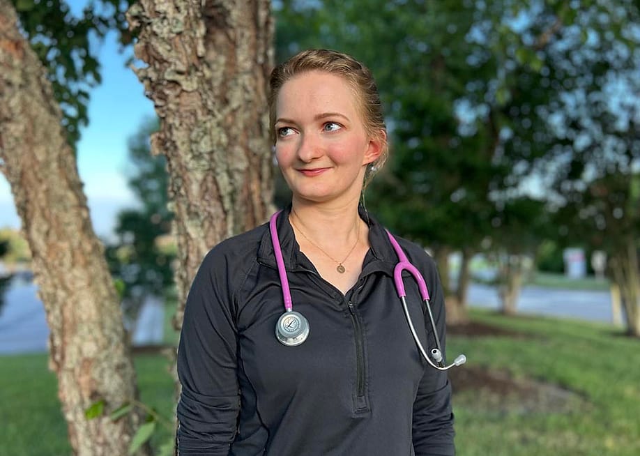 Michaela Bonner, wearing a black zip-up top with a pink stethoscope around her neck, poses for a portrait in front of some trees at local hospital in Virginia Beach, VA. — Jnaiah Price