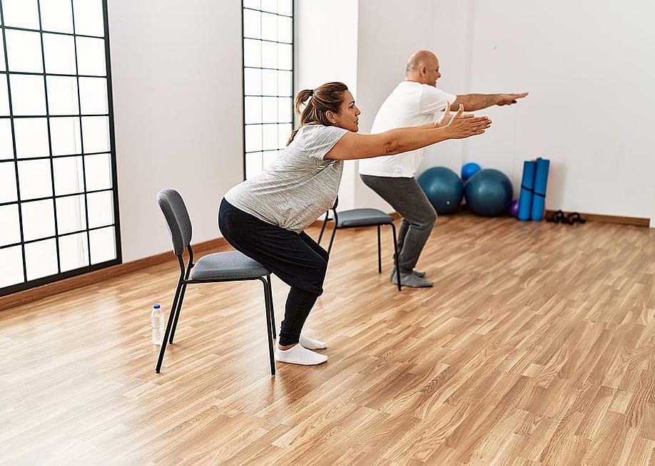 Two people each squatting toward a chair with their arms out in front of them, in a yoga studio type room. — Krakenimages.com // Shutterstock