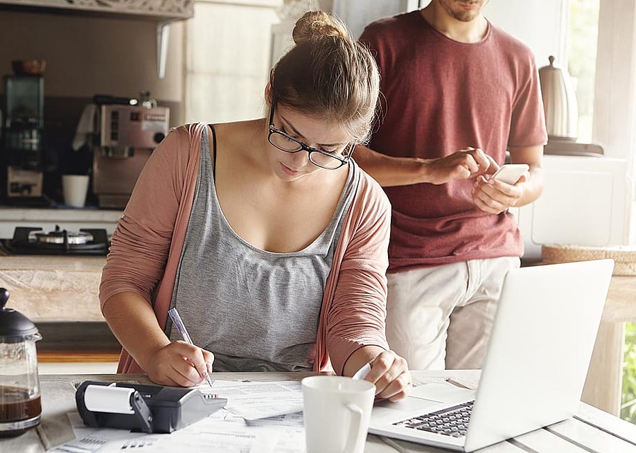 Woman doing calculations at kitchen table with laptop open and pen and paper while man with face not visible in the background is on his phone. — Cast Of Thousands // Shutterstock