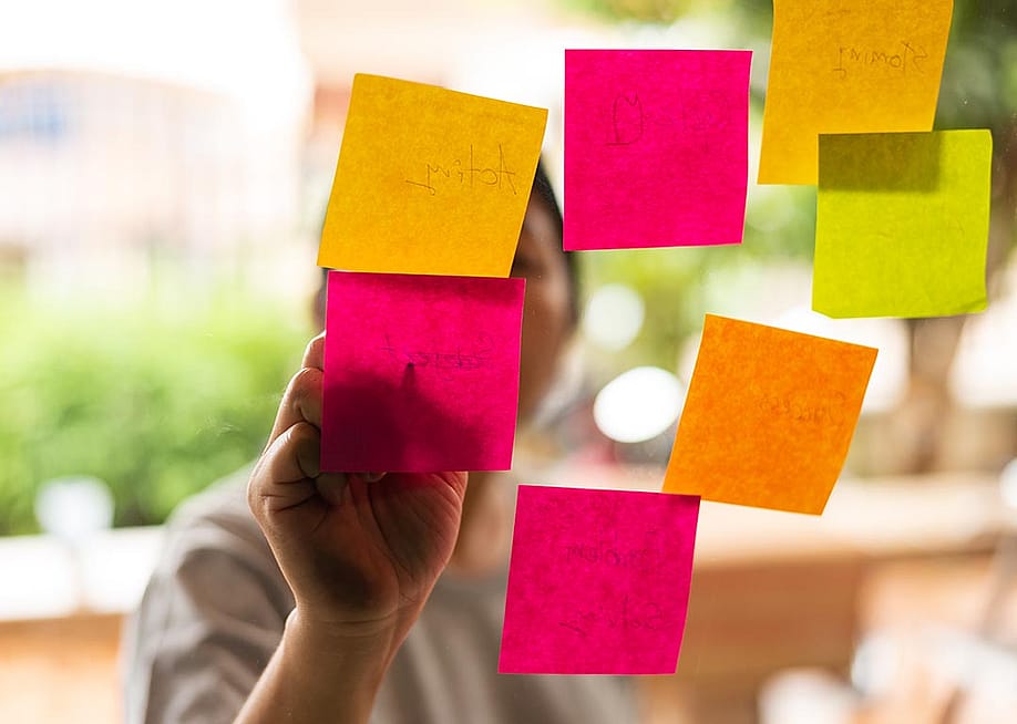 7 Pink, orange and yellow post-it notes stuck on glass with person whose face is obscured, writing on them from the other side. — Komsan Loonprom // Shutterstock