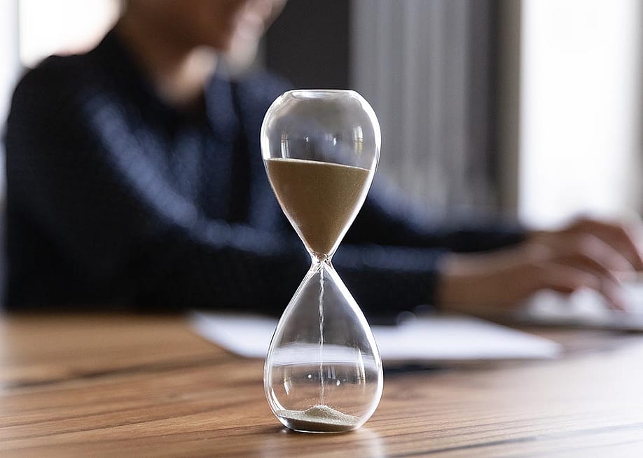 Close up view of hourglass stand on wooden office table measuring time, person working on laptop in the background blurred. — fizkes // Shutterstock