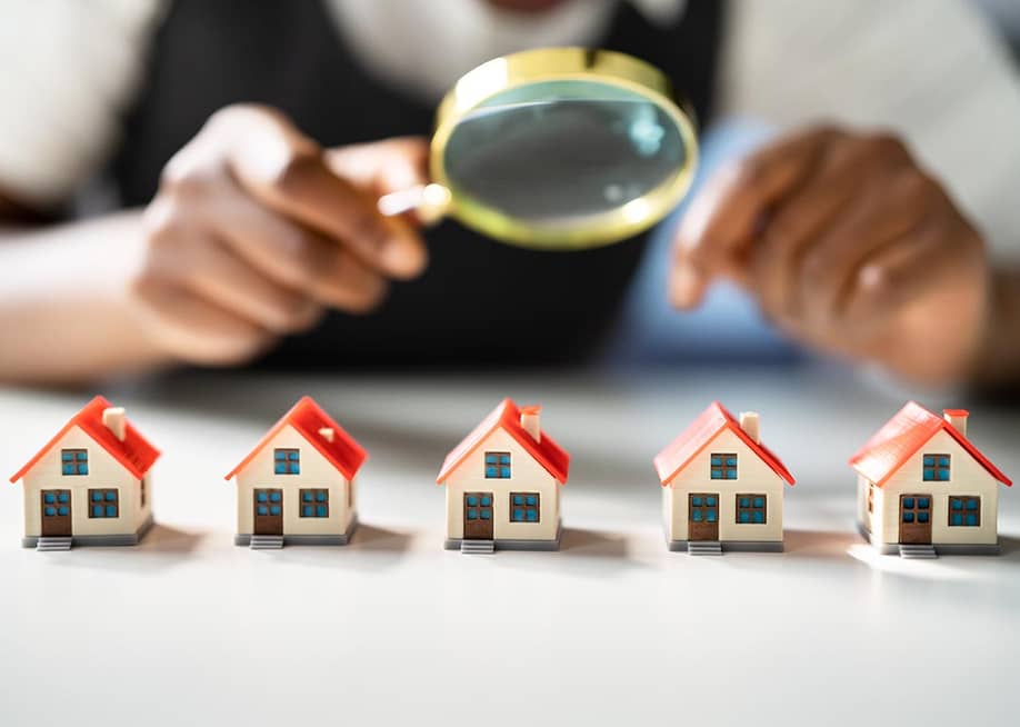 Five miniature houses with red roofs lined up on table, with magnifying glass held above them by a person symbolizing home and neighborhood appraisals. — Andrey_Popov // Shutterstock