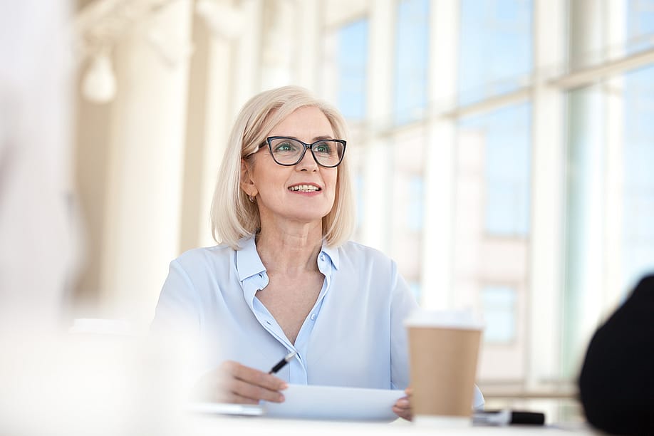 Blond professional sitting at table with large windows in the background talking in meeting. — fizkes // Shutterstock