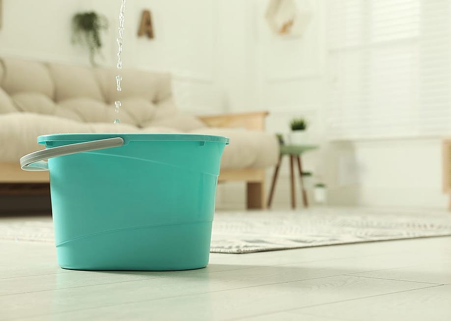 Close up of turquoise-colored plastic bucket on floor in living room with water dripping from ceiling into it. — New Africa // Shutterstock