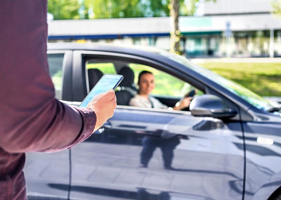 Arm of person in focus in the foreground, holding smartphone, with blurred dark car pulling up in the background, driver visible through the passenger window. — Tero Vesalainen // Shutterstock
