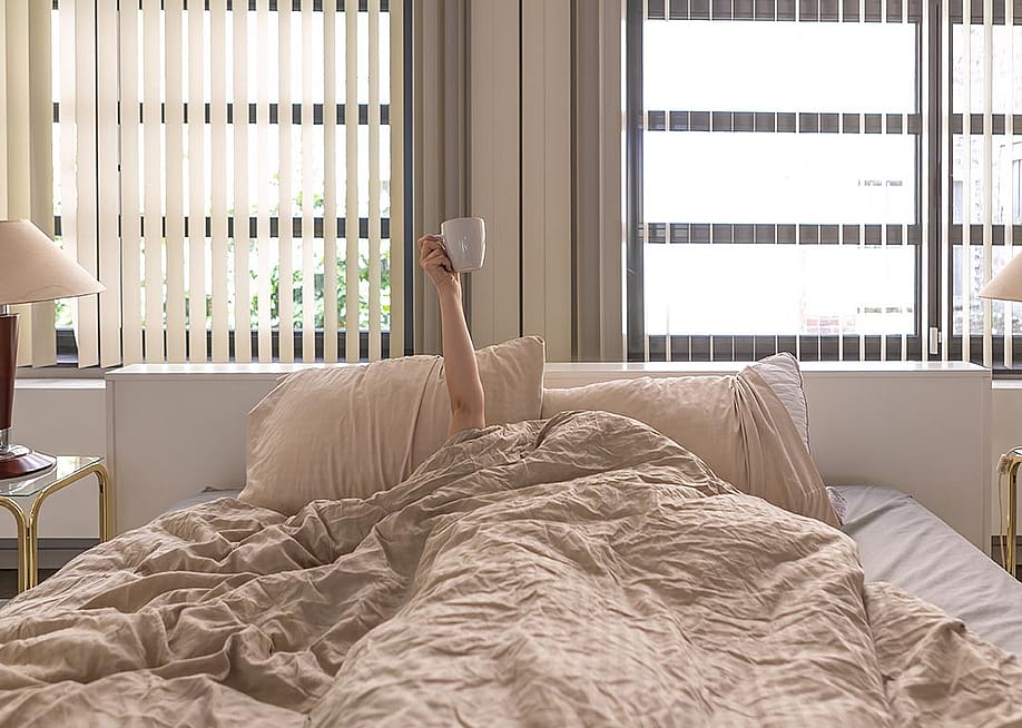 Hand holding a coffee cup extending upward from person whose face and body are under the covers in bed. — Carlo Prearo // Shutterstock