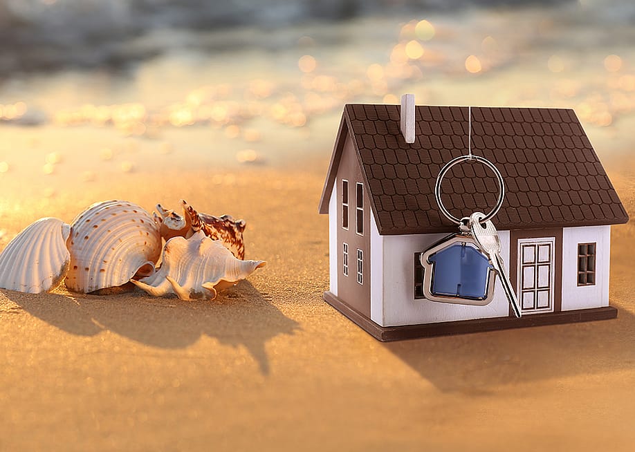 A model of a house with keys on a sandy beach beside seashells. — Pixel-Shot // Shutterstock