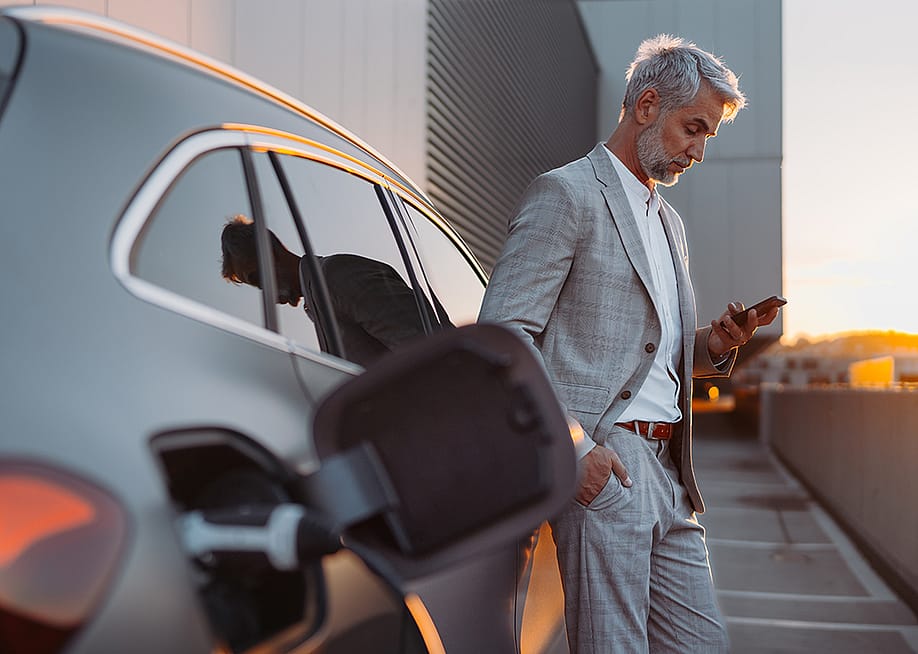 A man leaning on his electric car while using his smartphone. — Halfpoint // Shutterstock