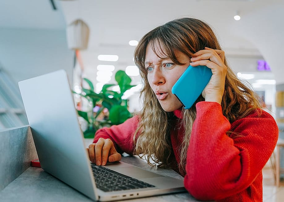 A woman talking on her smartphone while also booking flights using her laptop. — ShishkinStudio // Shutterstock