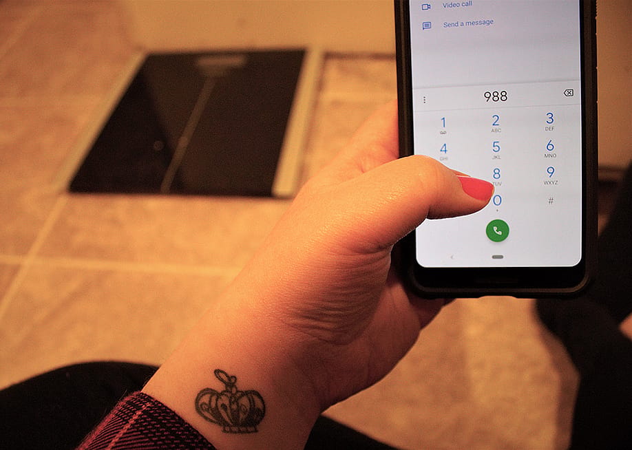 A woman with a tattoo dials the national suicide hotline '988' number from the floor in front of a body weight scale. — Lisa Schulz // Shutterstock