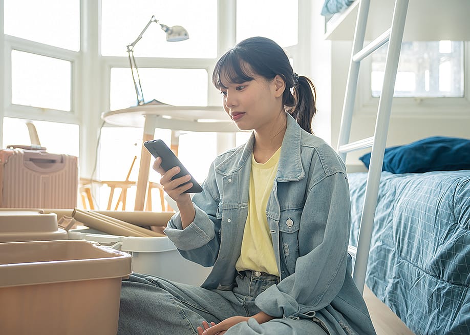 A college student using her smartphone in her dorm while unpacking personal items. — Lee Charlie // Shutterstock