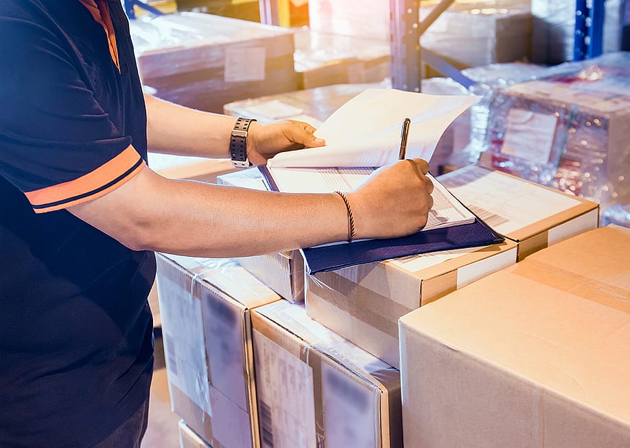 Warehouse worker writing on a clipboard on top of boxes as part of checking inventory. — Siwakorn1933 // Shutterstock