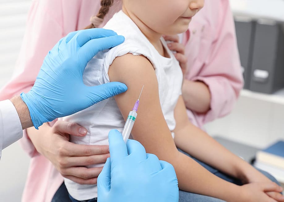 closeup of child's arm with vaccine needle held next to it by a person with blue gloves, an adult in a pink shirt holds child from other side. — New Africa // Shutterstock